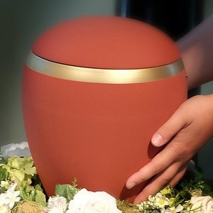 Rote Urne auf Blumenkranz von Händen umfasst